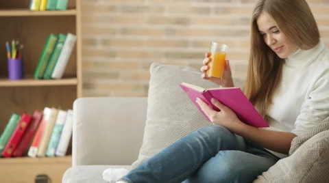 Charming girl sitting on sofa reading book drinking juice looking at camera  Stock Footage 34114586