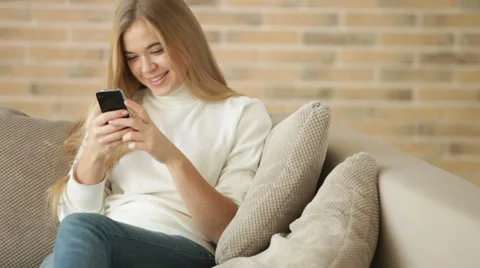 Charming girl sitting on sofa using mobile phone and smiling. Panning camera Video stock 34114533