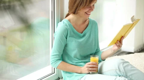 Charming girl sitting by window drinking juice reading book looking at camera Stock Footage 33851241
