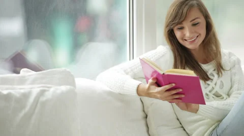 Charming girl sitting by window reading book closing it looking at camera  Stock Footage 34150141