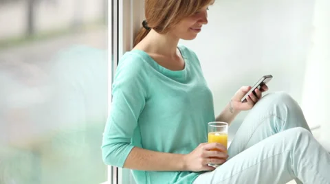 Charming girl sitting by window using cellphone drinking juice and smiling Stock Footage 33851128