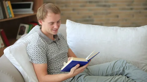 Charming guy relaxing on couch reading book looking at camera and smiling Stock Footage 34278049
