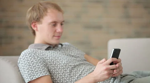 Charming guy relaxing on sofa using mobile phone looking at camera and smiling Stock Footage 34278035