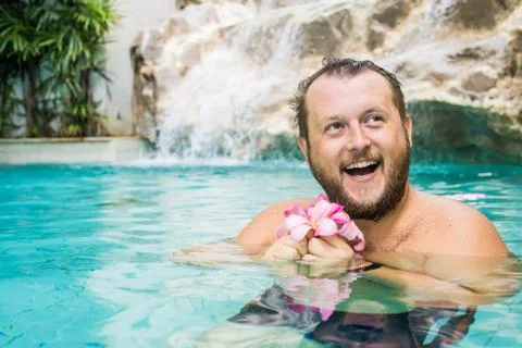 Charming man with a beard in a pool with blue water, holds a plumeria flower in Stock Photos