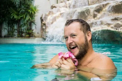 Charming man with a beard in a pool with blue water, holds a plumeria flower in Stock Photos