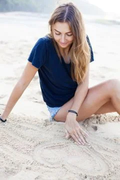 Charming woman sits on white beach and draws heart on sand. Stock Photos