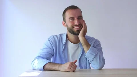 Charming young man with a beard sitting at a table and smiling while looking at Vídeos de archivo 344757289