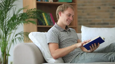 Charming  young man relaxing on sofa reading book looking at camera and smili Видео 34278056