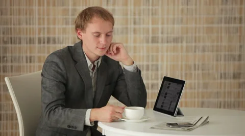 Charming young man sitting at table with touchpad drinking tea and smiling  Stock Footage 34278073
