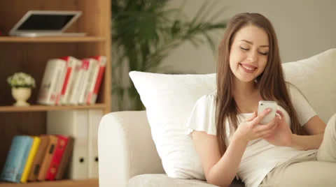 Charming young woman on sofa using mobile phone and smiling at camera Stock Footage 27177648
