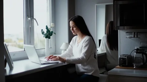 Charming young woman working behind a laptop next to a window in the kitchen. Stock Footage 119289341