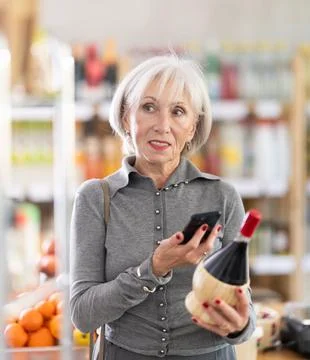 Checking expiration date of wine - elderly female shopper scans QR code on label 스톡 사진
