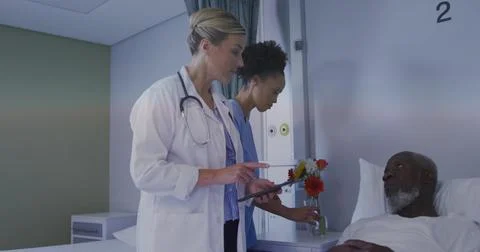 Checking lab coat doctor holding tablet at bedside, blue scrubs nurse placing Stock Photos