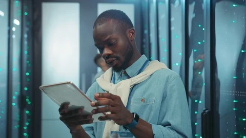 Cheerful african IT engineer browsing tablet news in server room calling for Stock Footage 119843393