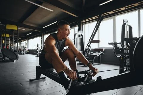 Cheerful attractive man smiling while using rowing machine in contemporary gym Stock Photos