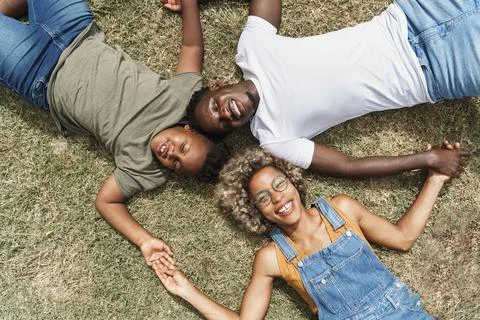 Cheerful Black family holding hands while lying on grass together in park. Happy Stock Photos