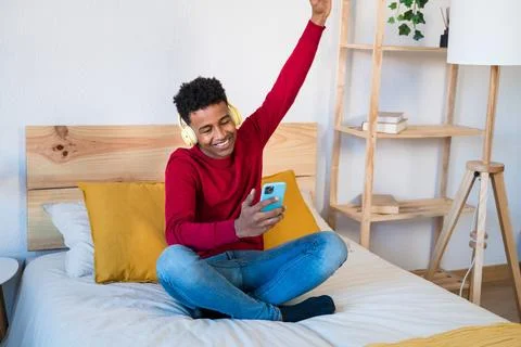 Cheerful black man using smartphone on bed listening music and dancing Stock Photos