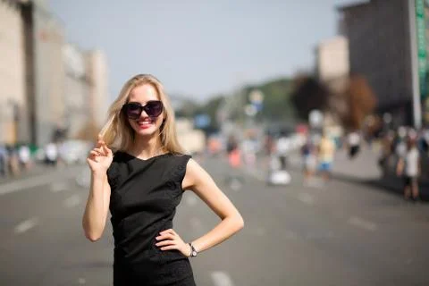 Cheerful blonde model wearing black dress and sunglasses, walking down the su Stock Photos