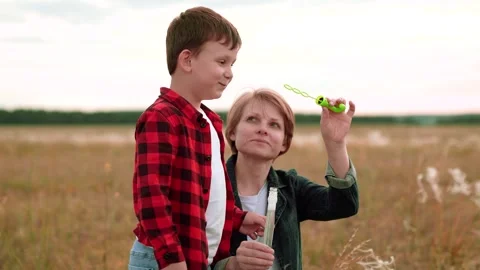 Cheerful boy in checkered shirt blows bubbles while mother helps on mowed field Stock Footage 256066183