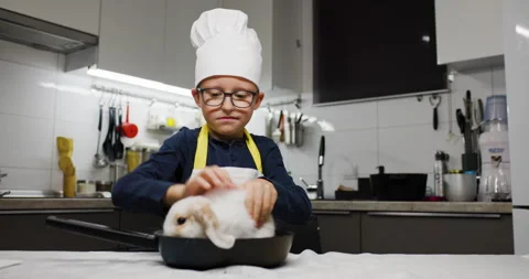 A cheerful boy in a chefs hat playing with a rabbit in a pan in a modern kitchen Stock Footage 321988846