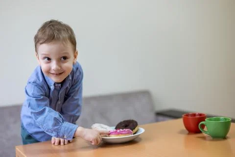 Cheerful boy reaching for donuts on table. Smiling young boy in blue shirt Stock Photos
