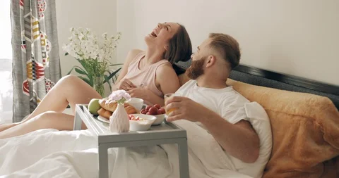 Cheerful couple having breakfast in bed and laughing while looking at each other Video stock 130043392