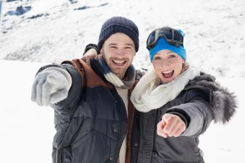 Cheerful couple in jackets pointing at camera on snow covered landscape Foto stock