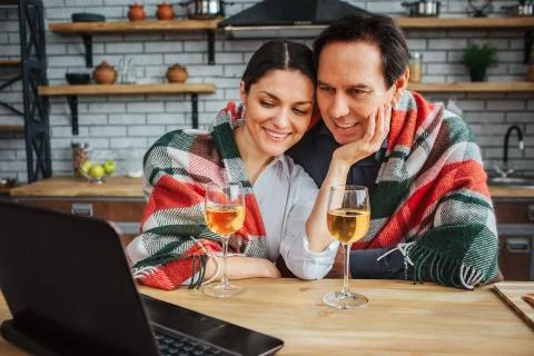 Cheerful couple sit at table in kitchen. Their shoulders covered with blanket 库存照片