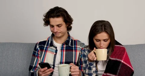 Cheerful couple sitting on sofa, drinking tea and using smartphone 스톡 동영상 125614750