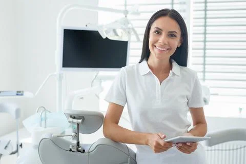 Cheerful dentist in coat using tablet in office and looking at the camera Stock Photos
