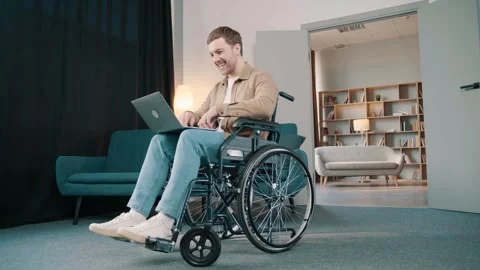 Cheerful disabled man on wheelchair using laptop in living room at home Video stock 244653297
