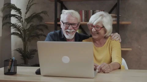 A cheerful elderly couple is web surfing the laptop indoors, a grey haired man Stock Footage 140868621