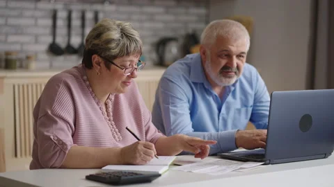 Cheerful elderly spouses are sitting together at kitchen and calculating utility Stock Footage 153399003