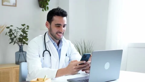Cheerful ethnic doctor using smartphone while working on laptop at home Stock Footage 228946595