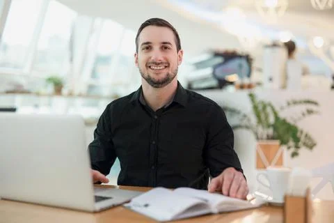 Cheerful freelancer man working from cafe. Attractive young man smiling, using Stock Photos