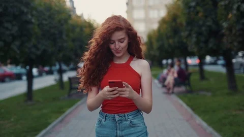 Cheerful ginger curly long haired student girl walks down the alley, uses Stock Footage 115669877