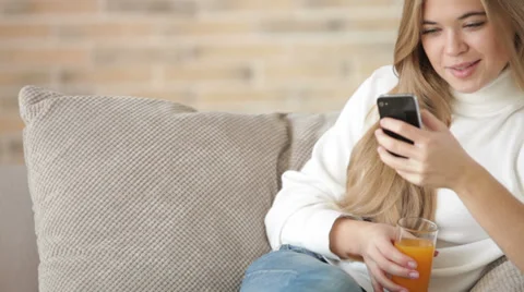 Cheerful girl sitting on sofa using mobile phone drinking juice and smiling.  Stock Footage 34115363