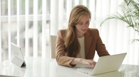 Cheerful girl sitting at table using touchpad and laptop looking at camera  Stock Footage 34835558