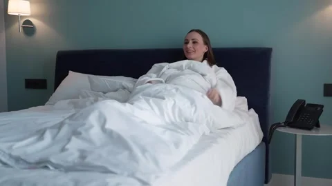 Cheerful girl in a white robe is lying in a comfortable bed in a hotel and Stock Footage 252659853