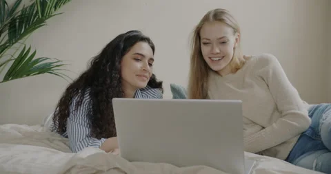 Cheerful girls using laptop to make online video call lying in bed and talking Video stock 236542985