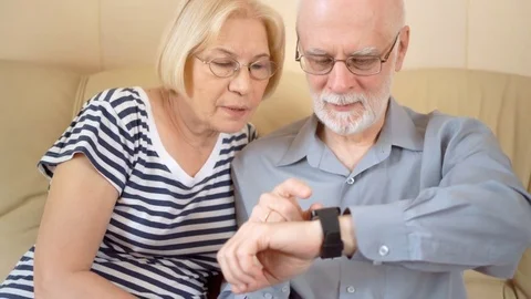 Cheerful good-looking senior couple sitting on sofa at home. Using smartwatch Stock Footage