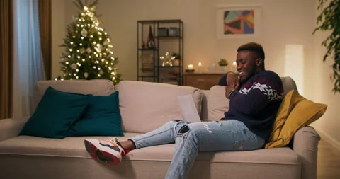 Cheerful guy of African appearance sits on couch and watches comedy movie on Stock Footage 211133393