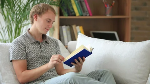 Cheerful guy relaxing on couch reading book looking at camera and smiling Stock Footage 34278121