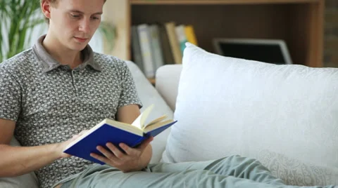 Cheerful guy sitting on sofa reading book looking at camera and smiling. Panning Stock Footage 34278163