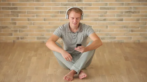 Cheerful guy wearing headset sitting on floor using mobile phone and smiling  Vídeos de archivo 34278193