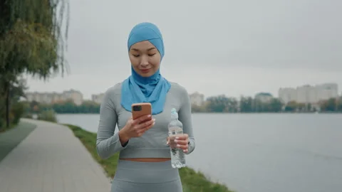 Cheerful Hijab Lady Using Smartphone and Holding Water from Plastic Bottle Stock Footage 259688441
