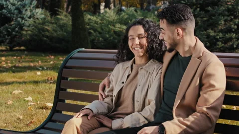Cheerful hispanic couple cutely communicate sitting on bench in autumn city park Stock Footage 165833745