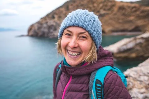 A cheerful, joyful blonde in a knitted hat laughs, standing on the seashore.  Stock Photos