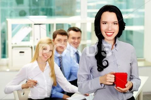 Cheerful lady holding a cup during coffee break, her colleagues smiling from beh Stock Photos