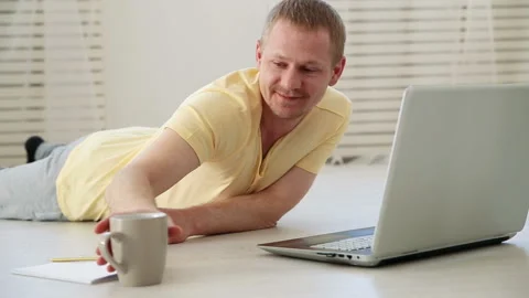 Cheerful man drinking coffee and chatting on laptop at home on the floor Stock Footage 91257475